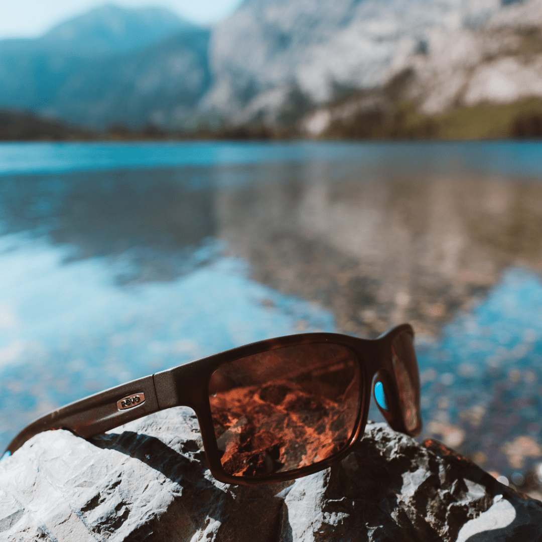 Sunglasses with tortoiseshell pattern on a rock by a lake with mountains in the background.