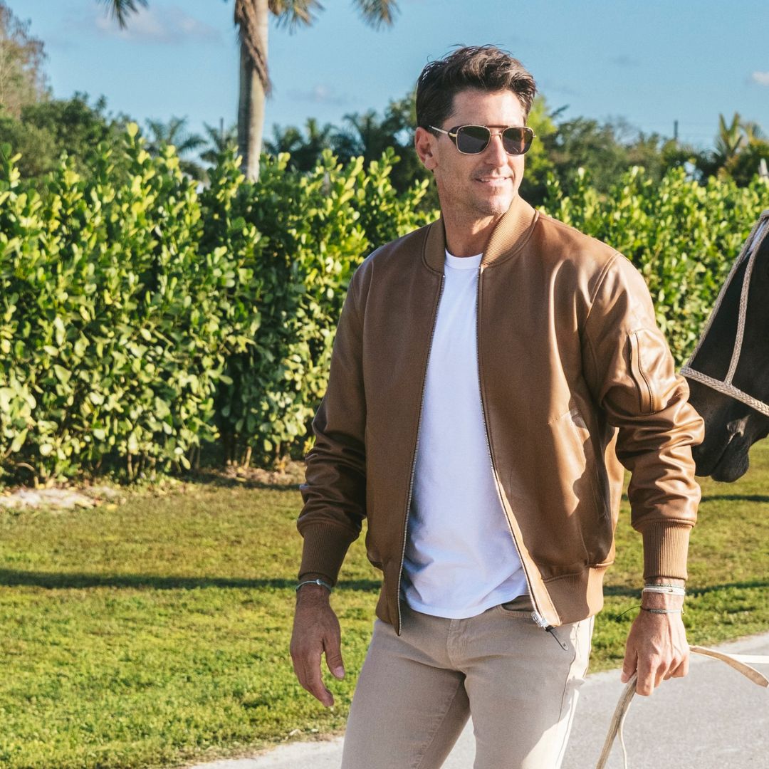 Man in brown jacket and sunglasses walking outdoors with a horse in the background