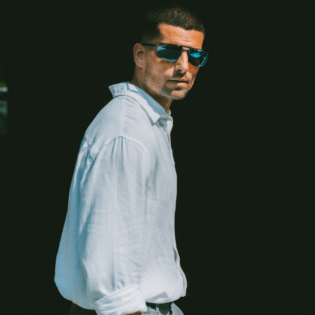 Man wearing sunglasses and a light-colored shirt against a dark background
