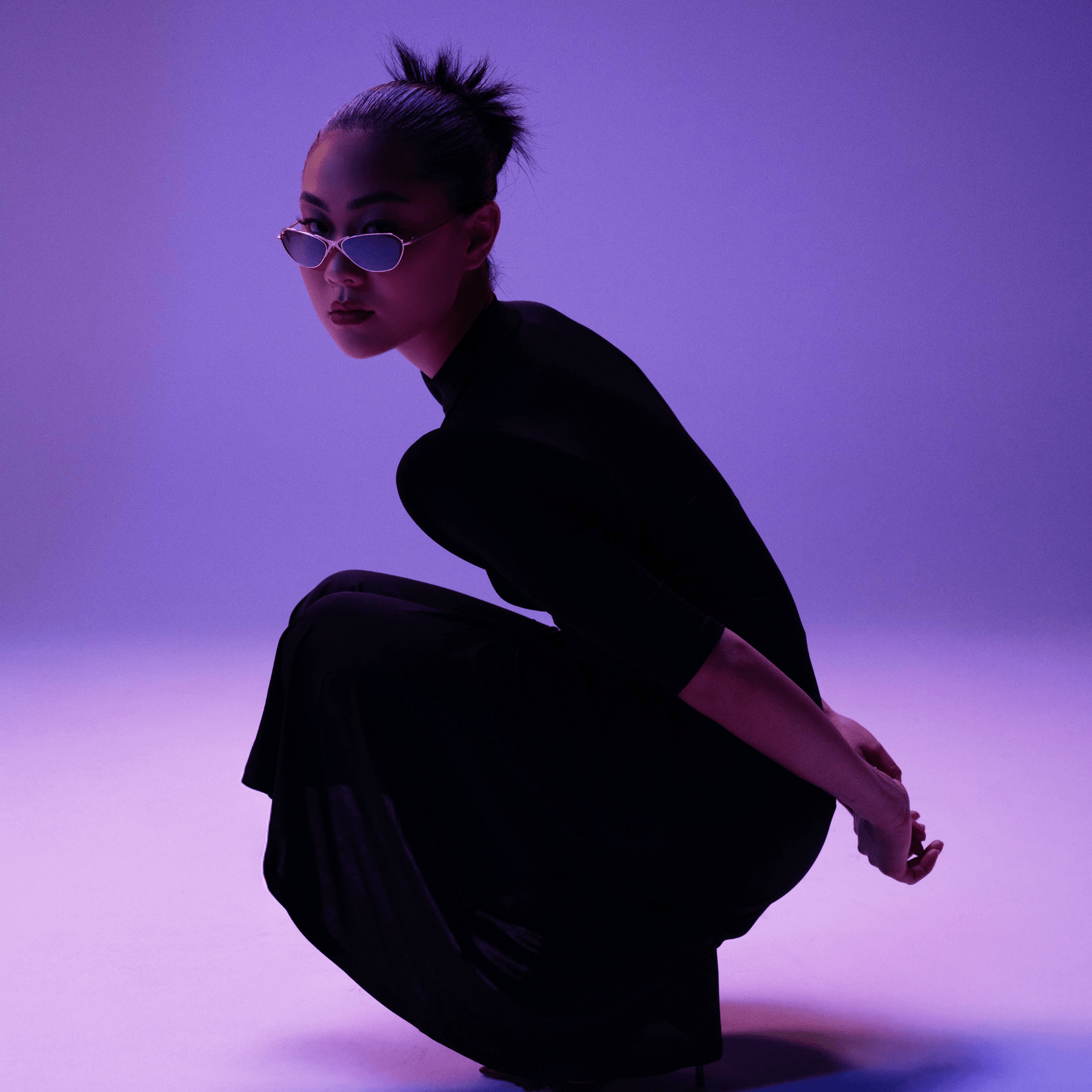Person in black outfit and sunglasses crouching against a purple background