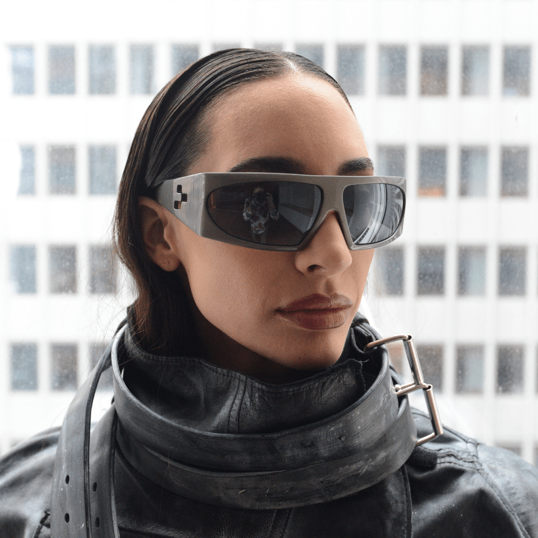 Person wearing futuristic sunglasses and a high-collared outfit with a blurred cityscape background