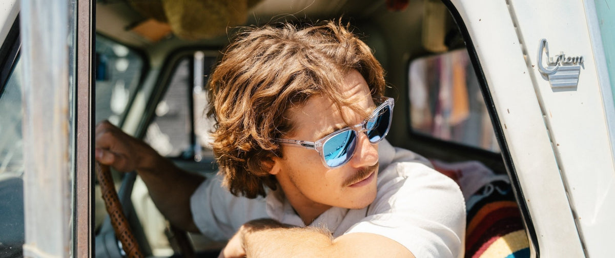 Man with tousled hair in a vintage white car wearing clear frame sunglasses with blue lenses
