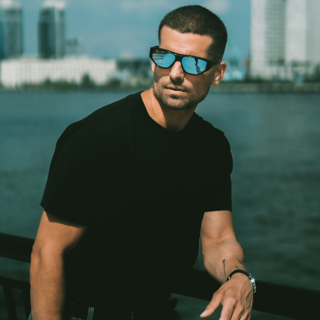 Man wearing sunglasses and a black t-shirt by a waterfront with buildings in the background