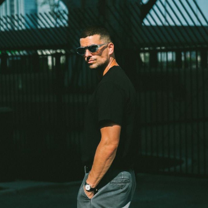 Man wearing sunglasses and a black shirt standing in a sunlit area with shadows.