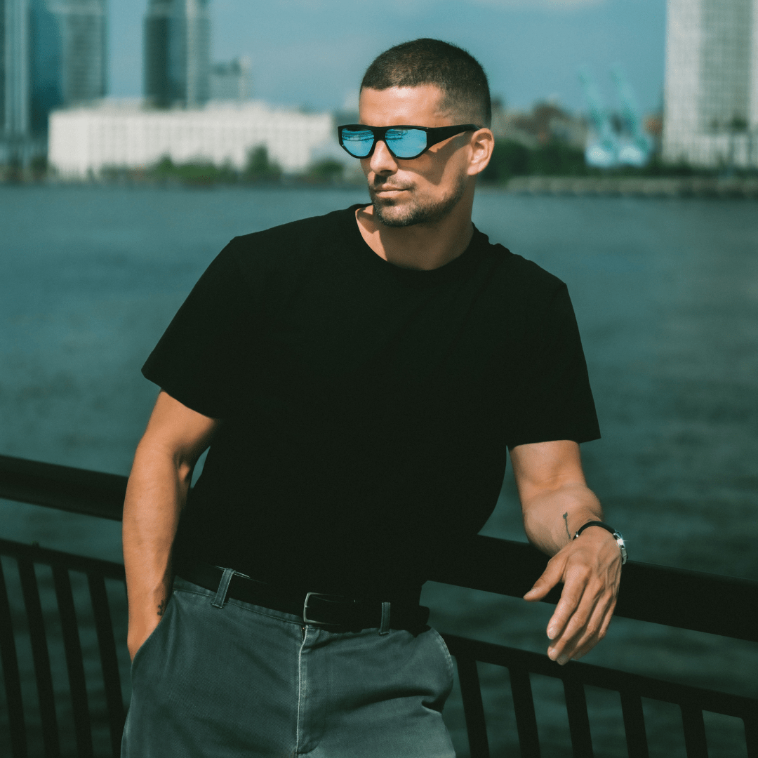 Man wearing sunglasses and a black t-shirt by a waterfront with city skyline in the background