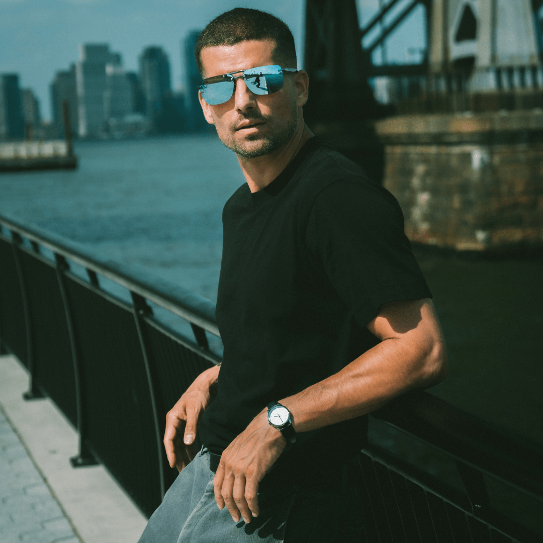 Man wearing sunglasses and a black t-shirt by a waterfront with a city skyline in the background