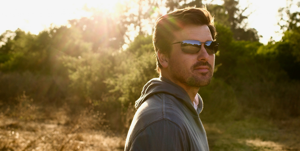 A man wearing dark mirrored sunglasses and a gray hoodie stands outdoors in warm golden sunlight with trees and brush in the background.