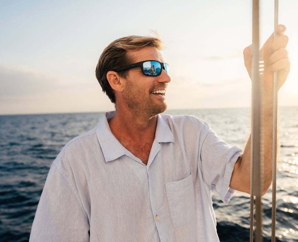 A smiling man wearing mirrored sunglasses and a light-colored button-up shirt stands on a boat with the ocean and sunset in the background.