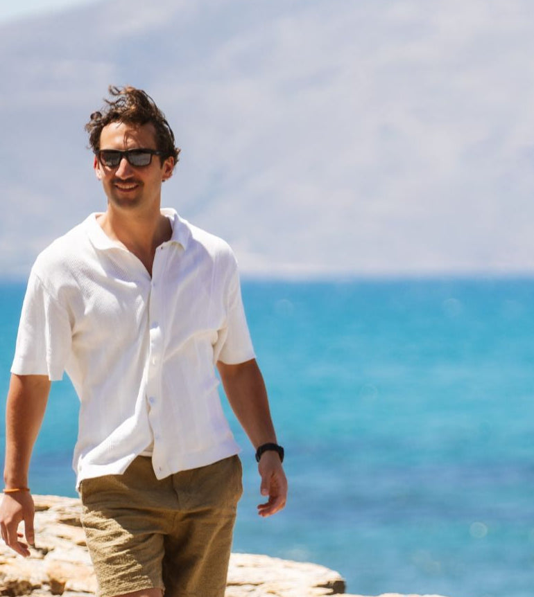 Man in a white shirt and shorts standing on a rocky outcrop with a scenic ocean view.