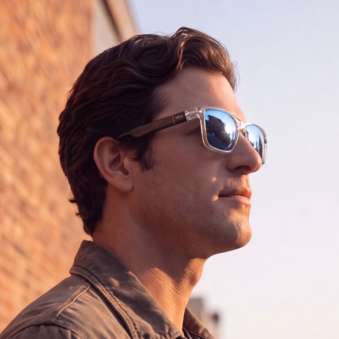 A man in profile wearing square sunglasses with a clear frame and mirrored blue lenses, photographed outdoors against a brick wall and pale sky.