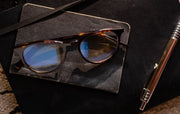 Round blue light glasses with computer screen reflection sitting on dark notebook with pen