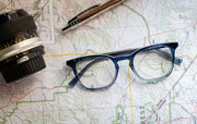 Vintage keyhole square blue light blocking glasses with clear blue frame sitting on map with pen and camera lens
